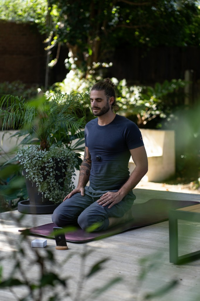 A person kneels in still meditation outdoors, eyes closed, small device on the chest, phone and stand nearby