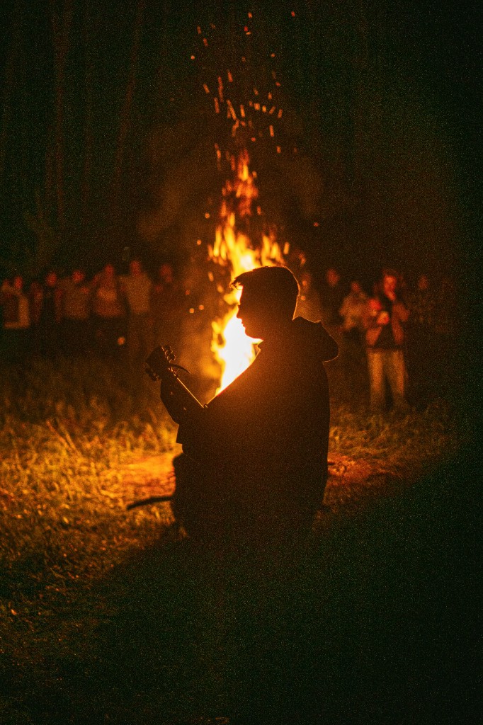 Night circle around a tall fire, sparks rising, someone plays guitar in silhouette
